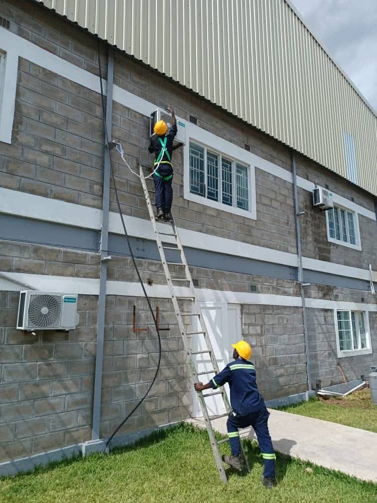 Harnessed technicians in hard hats installing air-conditioning units on a warehouse exterior