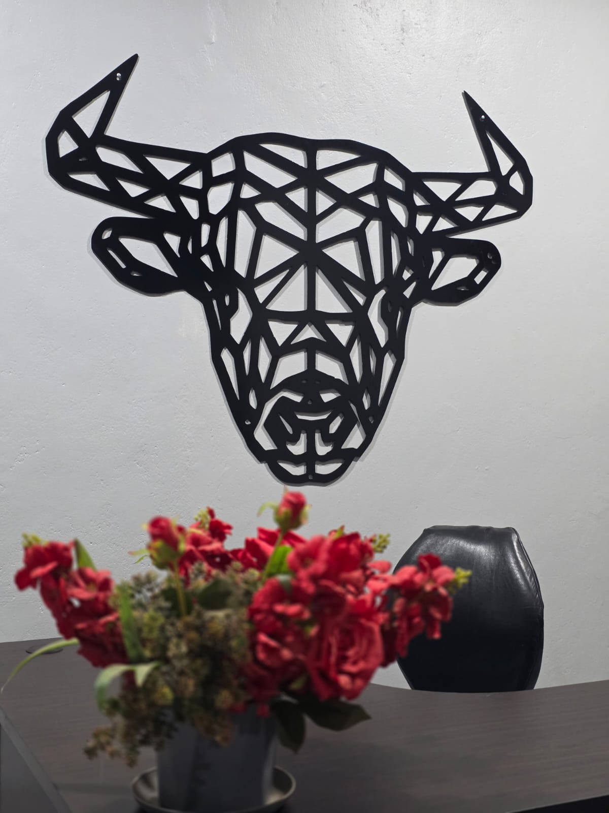 Black powder-coated geometric bull's-head steel wall art above a vase of red flowers